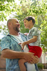 Happy father holding cute son with Down Syndrome below tree - CAIF33962