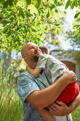 Happy father holding, hugging son under tree in summer backyard - CAIF33966