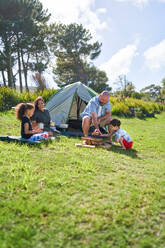 Happy family camping, stacking firewood outside tent in sunny grass - CAIF33980