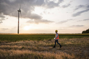 Engineer walking in field with wind turbine at sunset - UUF29269