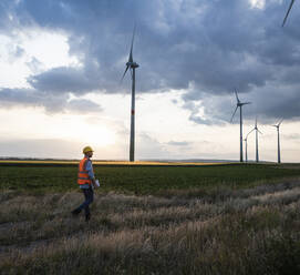 Engineer walking on wind field in front of sunset sky - UUF29270