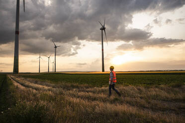 Engineer walking in field with wind turbines at sunset - UUF29271