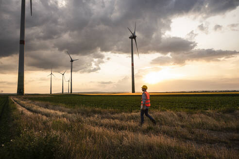 Ingenieur, der bei Sonnenuntergang in einem Feld mit Windturbinen spazieren geht - UUF29271