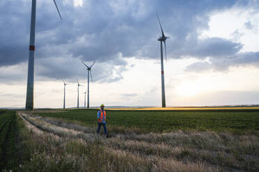 Engineer walking on agricultural field with wind turbines in front of sky - UUF29272