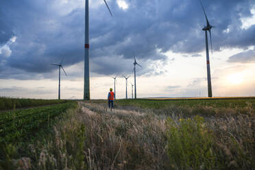 Engineer walking towards wind turbines in field at sunset - UUF29277