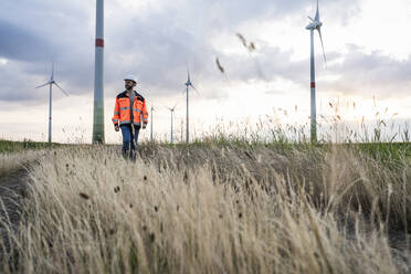 Engineer walking in grass with wind turbines in background - UUF29290