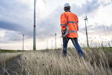 Engineer in reflective clothing walking with laptop at wind farm - UUF29292
