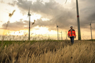 Engineer walking amidst grass on wind farm at sunset - UUF29293