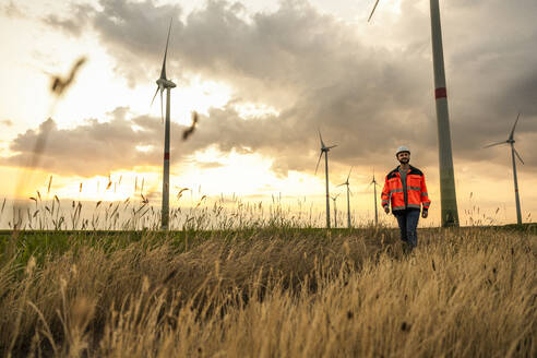 Ingenieur, der bei Sonnenuntergang in einem Windpark im Gras spazieren geht - UUF29293