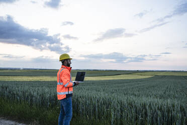 Ingenieur in reflektierender Kleidung, der einen Laptop hält und die Ernte auf einem Feld betrachtet - UUF29298