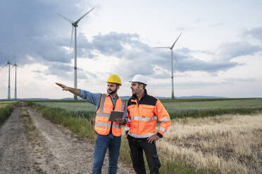 Engineer explaining colleague standing on footpath at wind farm - UUF29309