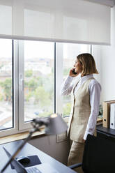 Geschäftsfrau, die mit einem Mobiltelefon spricht und durch ein Fenster im Büro schaut - EBSF03636