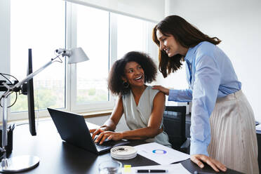 Smiling businesswoman discussing with colleague over laptop at desk in office - EBSF03642