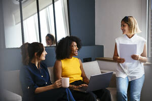 Glückliche multirassische Geschäftsfrauen, die auf dem Sofa sitzen und in einem kreativen Büro diskutieren - EBSF03652