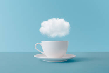 Cloud floating above white ceramic cup of aromatic drink representing concept of thoughts during coffee break on blue background - ADSF44407