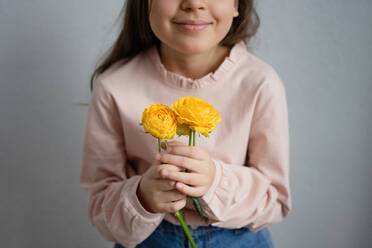 Crop young preteen female in casual clothes smiling and looking at camera while standing on gray background with yellow ranunculus flowers in hands - ADSF44430