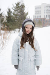 Positive girl in warm clothes smiling and looking at camera while standing in winter park against blurred green trees and buildings - ADSF44431