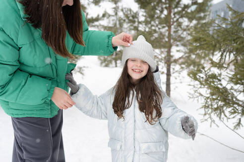 Cropped unrecognizable female in warm clothes adjusting cap of hugging preteen daughter and standing in winter park against blurred trees - ADSF44433