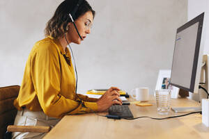 Telemarketing agent making calls and talking to potential customers through her headset. Woman sitting at her desk in the office, using a computer for her marketing work. - JLPPF02079