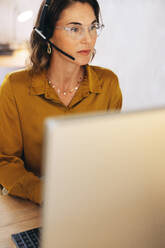 Woman working as a virtual assistant in a remote office. She is sitting at her desk, wearing a headset and using a computer as she provides client support over phone calls. - JLPPF02080