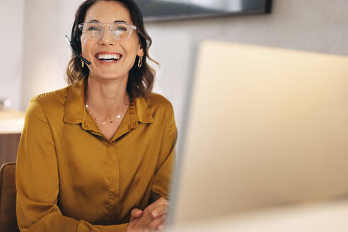 Happy travel agent sitting at her desk with a headset, assisting customers over phone calls. Woman using her communication skills to provide professional customer service for an online travel agency. - JLPPF02083