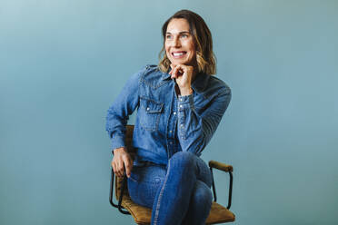 Female professional in her 30s, dressed in a denim jacket and business casual attire, sits in a chair in a studio. With a smile on her face, she looks away thoughtfully and contemplates her next move. - JLPPF02092