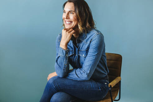 Woman in her 30s, an accomplished entrepreneur, radiates joy and determination in a studio environment. Dressed in business casual attire, including a denim jacket, she sits in a chair with a smile. - JLPPF02095