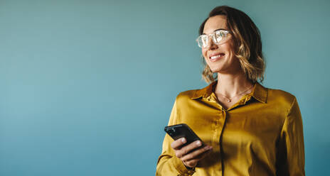 Caucasian business woman stands in a studio with a blue background, smiling and using her smartphone for work as an entrepreneur and freelancer. - JLPPF02098