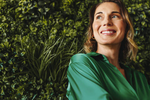 Woman smiling while contemplating creative ideas for launching an environmentally responsible business. Caucasian business professional standing in an office with a green interior design. - JLPPF02109