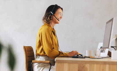 Happy call center agent sitting in front of a computer with a headset, answering calls and offering customer support. Woman assisting remote customers in a telecommunications job. - JLPPF02122