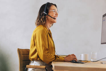 Happy tech support agent sitting at her workstation, wearing a headset and talking to calling customers. Woman providing customer service and expertly handling telecommunication tasks in a call centre. - JLPPF02124