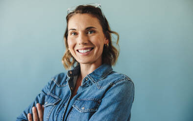 Portrait of an executive business woman smiling with confidence in a studio. Wearing a stylish denim jacket, she stands tall and looks directly at the camera with crossed arms. - JLPPF02138