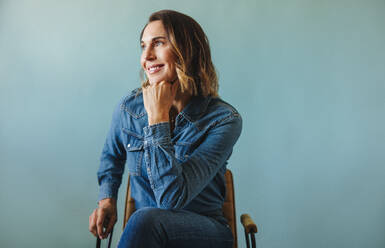 Female CEO and company owner looking away with a thoughtful expression in a studio. Business woman sitting on a chair in denim clothing, contemplating her next move in the business world. - JLPPF02147