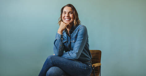 Successful business woman sitting on a chair in a studio, wearing a denim business attire. Professional woman celebrating her career as woman with experience and expertise in the business world. - JLPPF02148