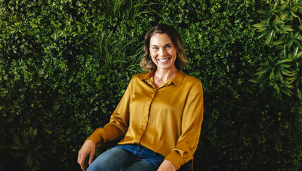 Businesswoman and CEO of a sustainable company sits in an office, with a green interior design in the background. Professional woman smiling at the camera; representing her company's eco-friendly values. - JLPPF02157