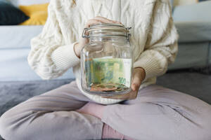 Woman holding money jar sitting cross-legged at home - TYF00801