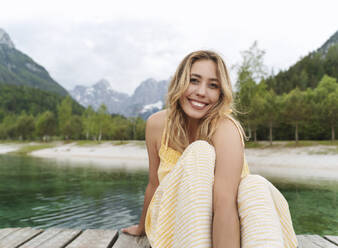 Beautiful smiling woman sitting on pier by river and mountains - NDEF00924