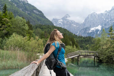 Backpacker with eyes closed relaxing on pier by mountains - NDEF00934