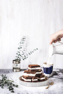 Die Hand einer Frau gießt Milch in eine Tasse mit Schokoladenkuchen auf dem Tisch - SBDF04621