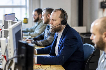 Mature businessman wearing headset sitting at desk with colleagues - WPEF07437