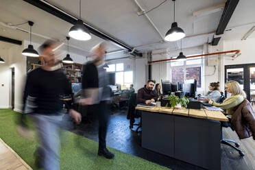 Businessmen walking with colleagues sitting at desk in office - WPEF07459