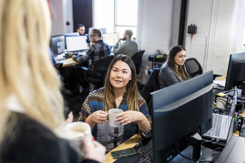 Geschäftsfrau mit Kaffeetasse im Gespräch mit einem Kollegen im Büro am Schreibtisch - WPEF07471