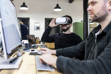 Happy businessman wearing virtual reality simulators with colleague working on computer in office - WPEF07493