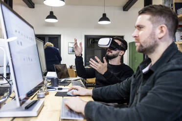 Businessman wearing virtual reality simulators with colleague working on desktop PC - WPEF07494