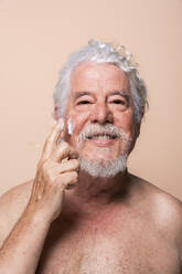 Älterer Mann, der eine Feuchtigkeitscreme auf die Haut aufträgt, vor beigem Hintergrund - OIPF03135