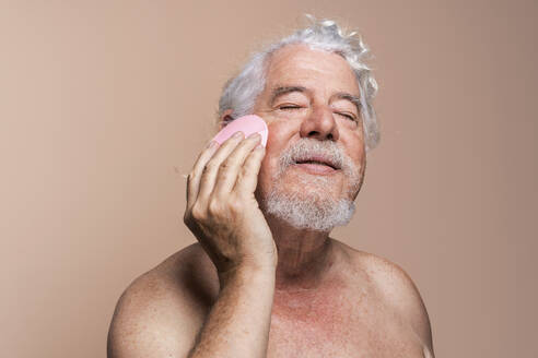 Älterer Mann mit geschlossenen Augen massiert sein Gesicht mit einem Pinsel vor beigem Hintergrund - OIPF03141