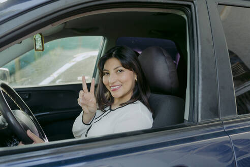 Happy woman showing peace sign gesture and sitting in car - DSIF00673