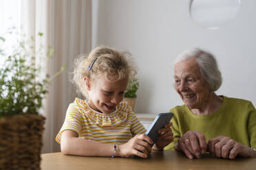 Smiling granddaughter using smart phone by grandmother at home - SVKF01504