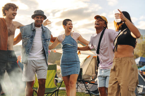 Group of happy young people having fun in the festive ambiance of a vibrant festival, drinking beer and celebrating summer life. Friends creating unforgettable memories at a camp. This photo has intentional use of 35mm film grain. - JLPPF02191