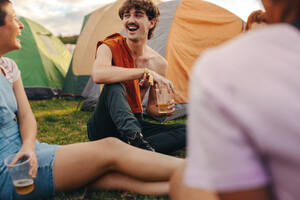 Group of friends gather outside their festival tents, creating a joyous scene, sharing laughter and drinking beers. Summer festival goers creating cherished memories together. This photo has intentional use of 35mm film grain. - JLPPF02196